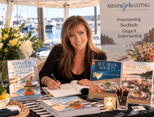 Missy Gervais smiling while signing a book at a waterfront boat show, surrounded by copies of Coastal Galley, Aft Deck Society and Teak Table, with the marina and Missy Goes Boating display in the background.
