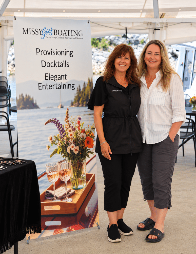 Missy Gervais and Tracy Sarich smiling beside a Missy Goes Boating banner at a marina book-signing event.